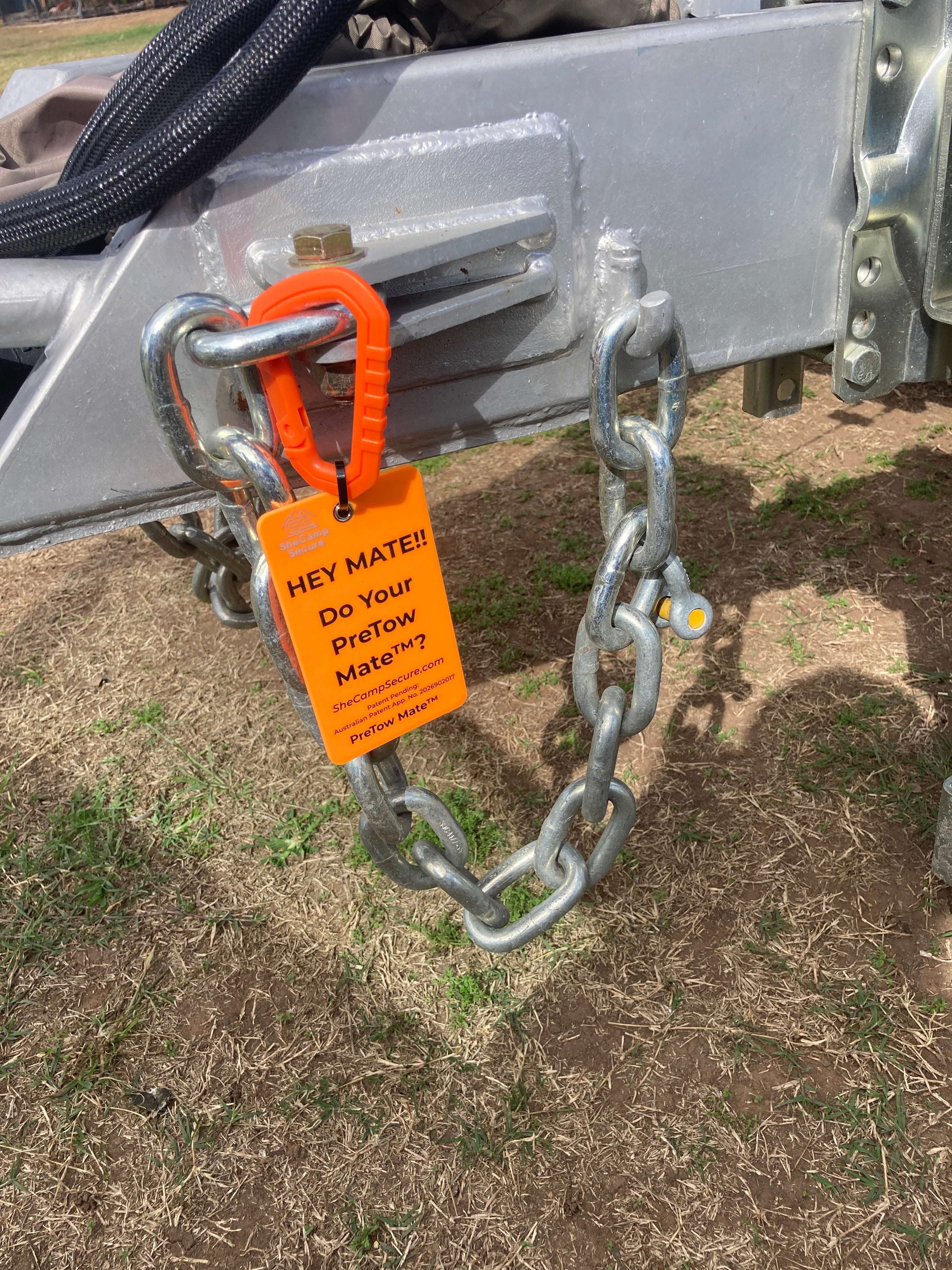 Close-up of an orange Hey Mate safety tag attached to a silver caravan chassis and safety chain, used as a pre-tow checklist reminder.