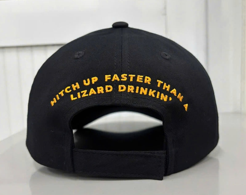 Rear view of the black baseball cap showing an adjustable fabric hook-and-loop strap closure. Arched across the back panels above the opening is the slogan "HITCH UP FASTER THAN A LIZARD DRINKIN'" embroidered in bold orange text.