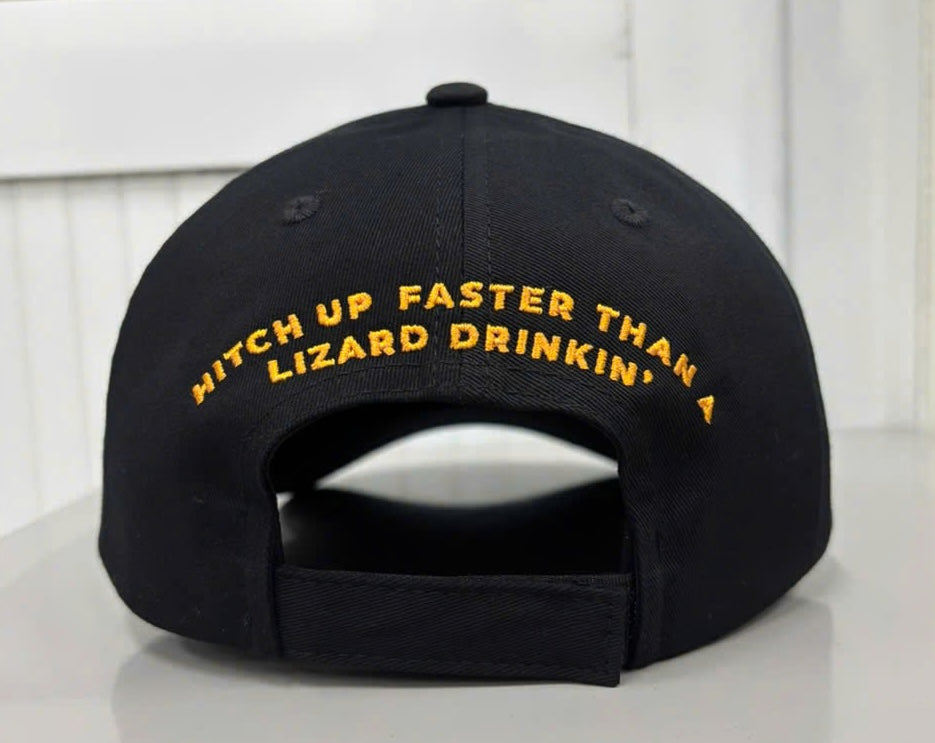 Rear view of the black baseball cap showing an adjustable fabric hook-and-loop strap closure. Arched across the back panels above the opening is the slogan "HITCH UP FASTER THAN A LIZARD DRINKIN'" embroidered in bold orange text.